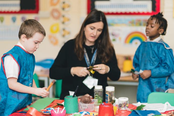 Image shows teacher helping two primary-aged children cut up coloured paper to make musical instruments
