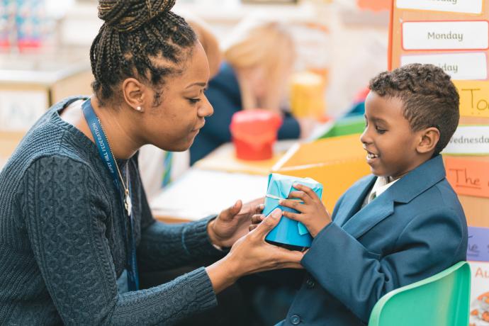 Image shows a teacher helping a primary aged pupil make a musical instrument out of paper and card